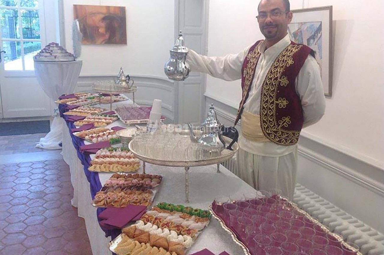 Un homme en tenue traditionnelle sert du thé près d'une table garnie de pâtisseries et de verres dans une salle lumineuse.