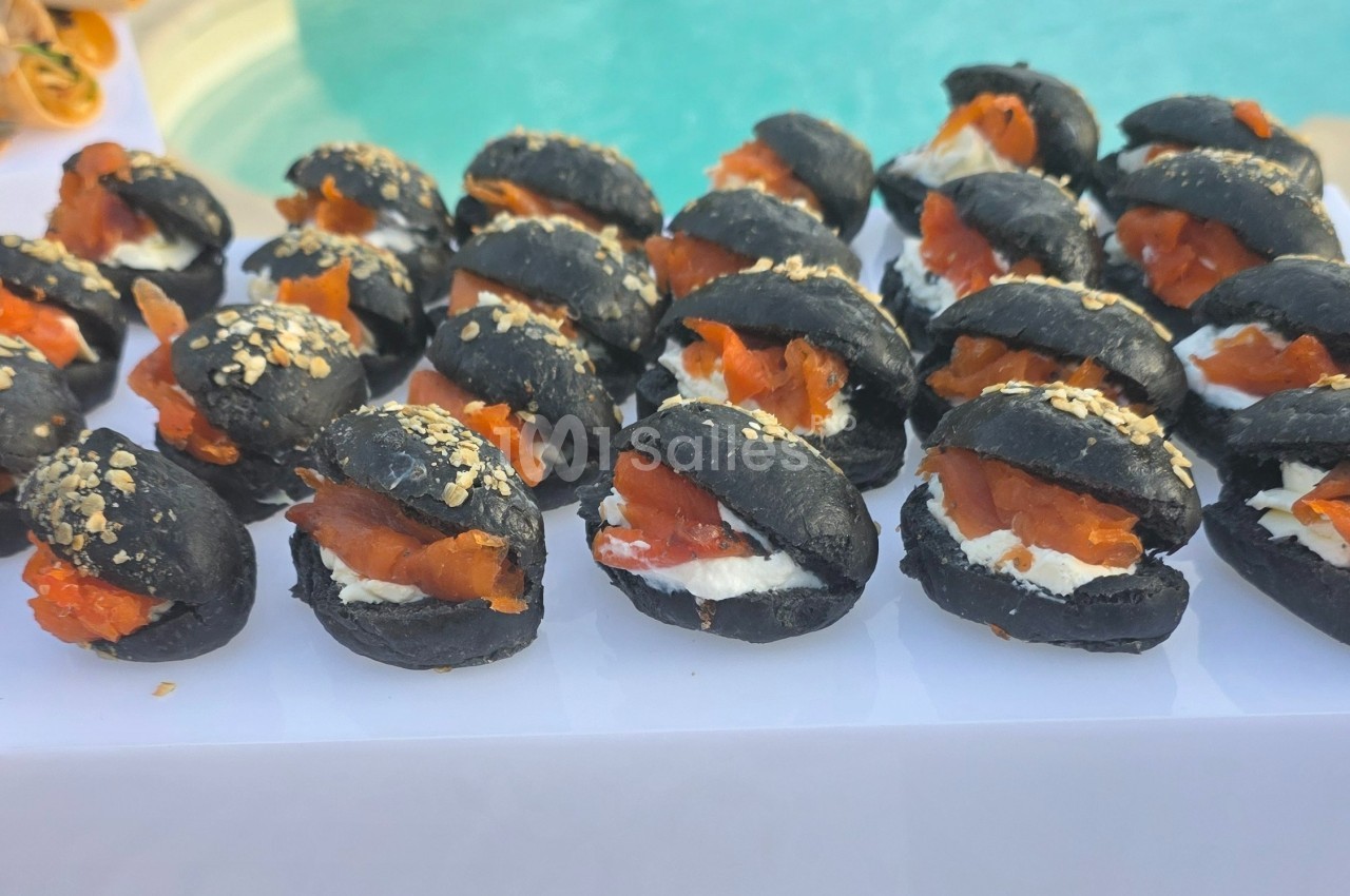 Petits pains noirs garnis de fromage frais et de saumon fumé, présentés sur un plateau près d'une piscine.