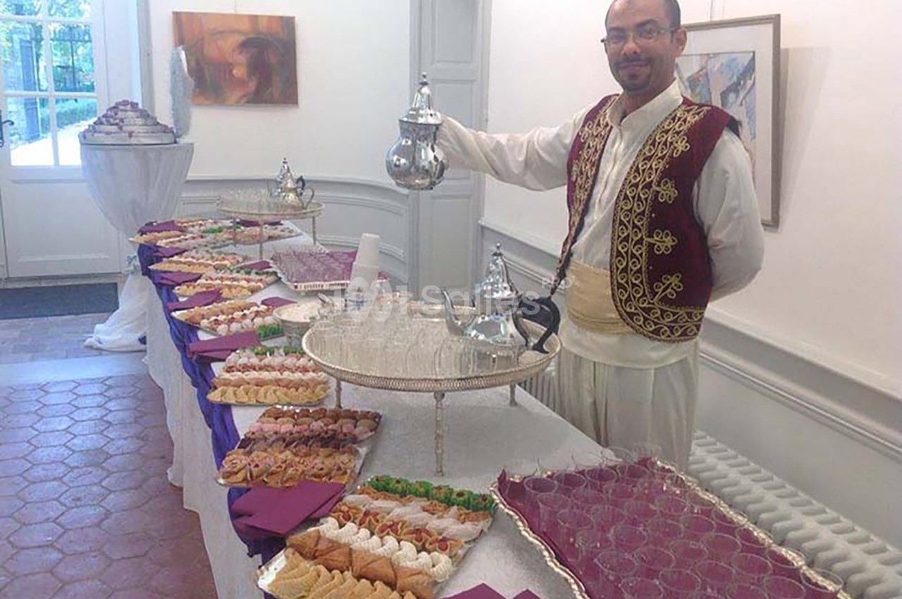 Un homme en tenue traditionnelle sert du thé près d'une table garnie de pâtisseries et de verres dans une salle lumineuse.