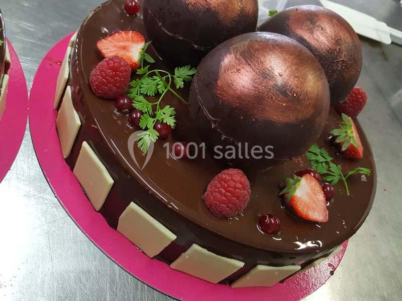 Gâteau au chocolat décoré de sphères brillantes, fruits rouges et carrés de chocolat blanc sur un plateau rose.