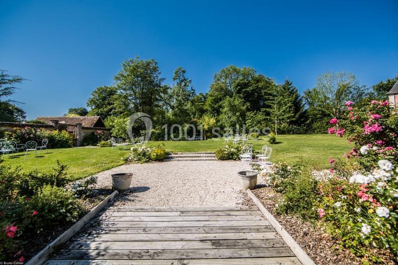 Jardin avec pelouse, arbres, mobilier de jardin blanc et allée en bois bordée de fleurs sous un ciel bleu.