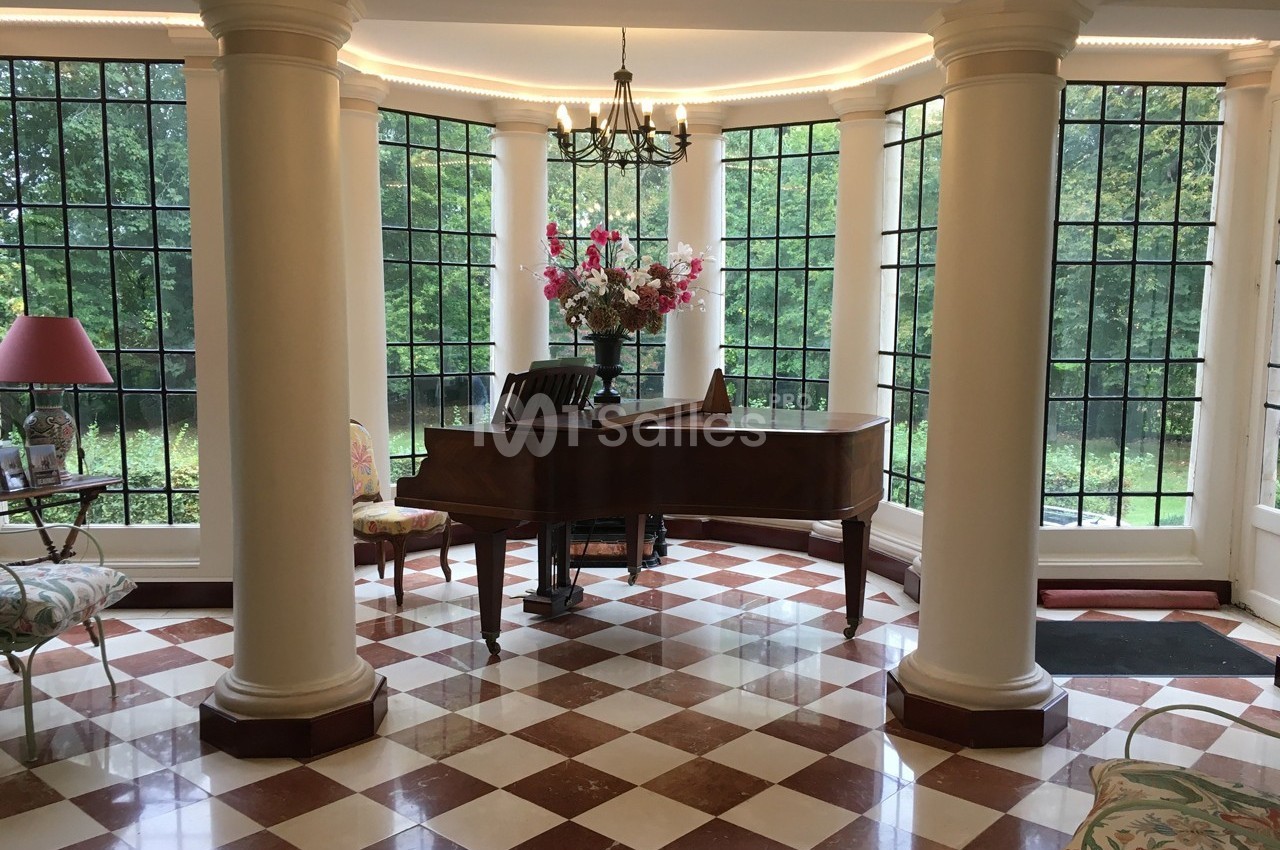 Salle lumineuse avec un piano à queue, des colonnes blanches, un sol en damier et de grandes fenêtres donnant sur un jardin.