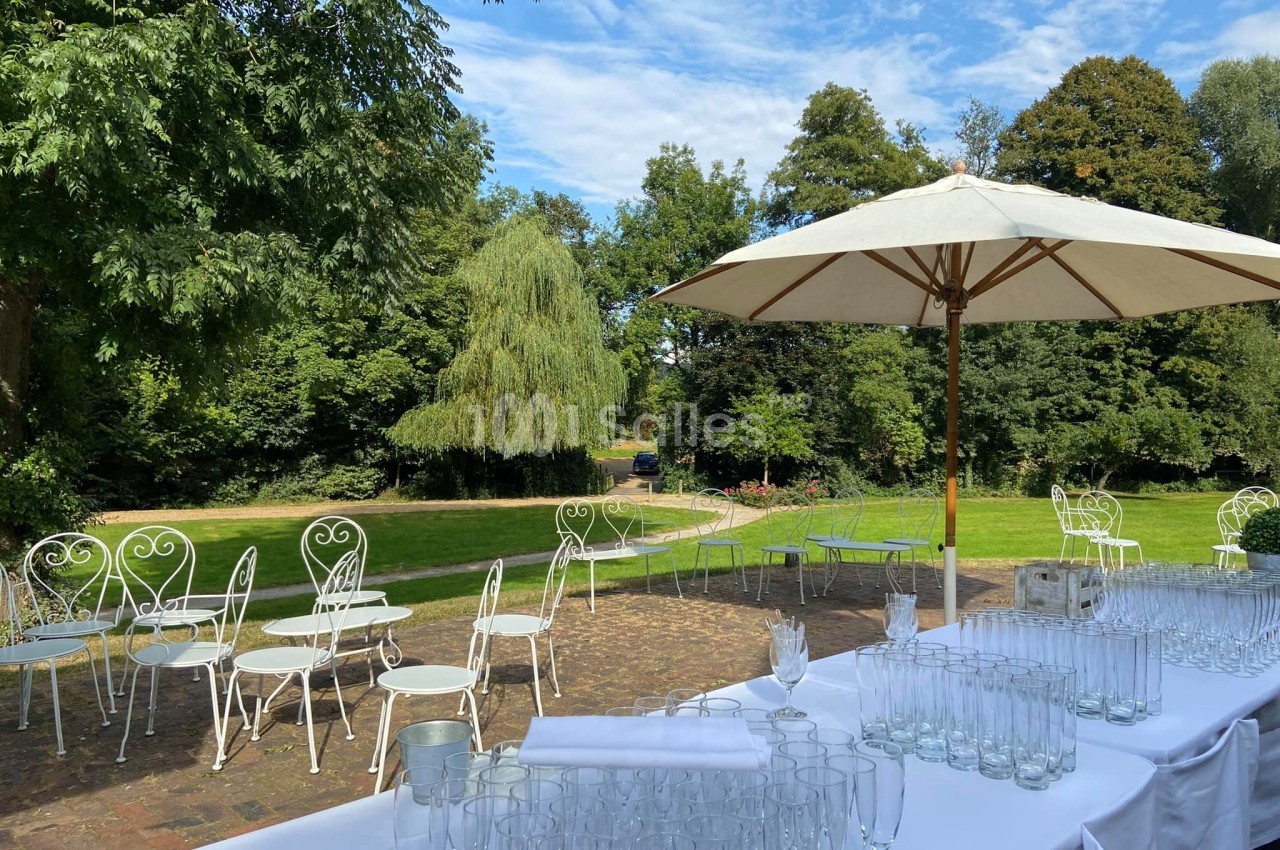 Tables et chaises blanches disposées sur une terrasse en pierre, avec un parasol et un jardin verdoyant en arrière-plan.