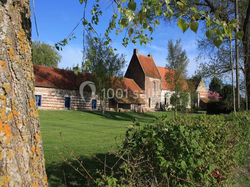 Bâtiment en briques et pierres avec toit en tuiles rouges, entouré d'arbres et d'une pelouse verdoyante.
