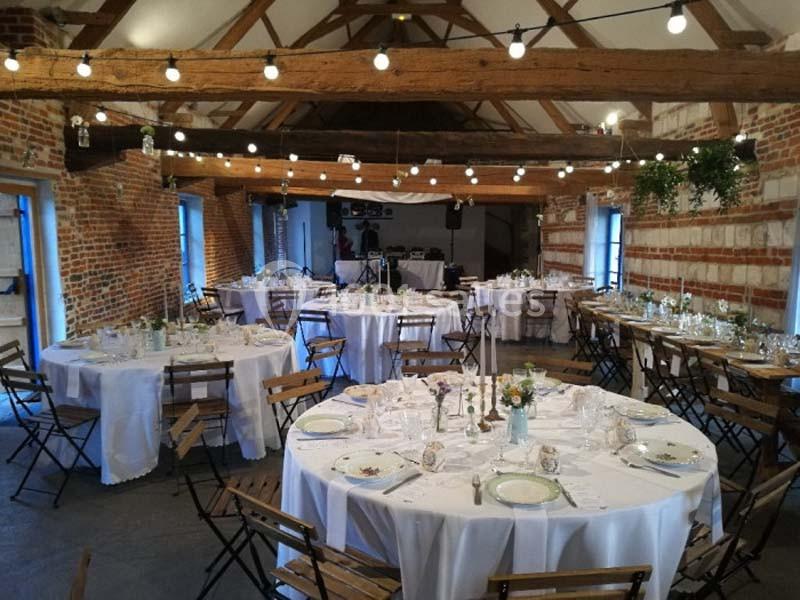Salle de réception rustique avec tables rondes dressées, chaises en bois et guirlandes lumineuses suspendues.