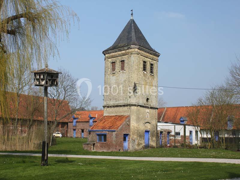 Tour en briques avec toit en ardoise, entourée de bâtiments ruraux et d'arbres, sous un ciel dégagé.