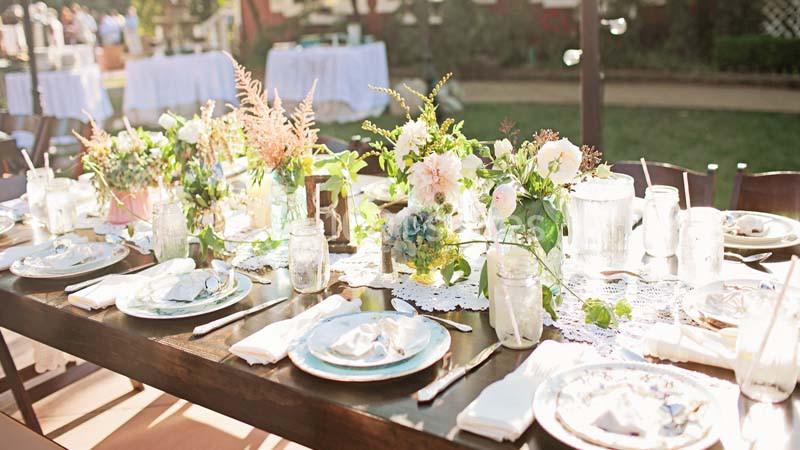 Table en bois décorée de fleurs, vaisselle blanche et nappes en dentelle, disposée pour un repas en extérieur.