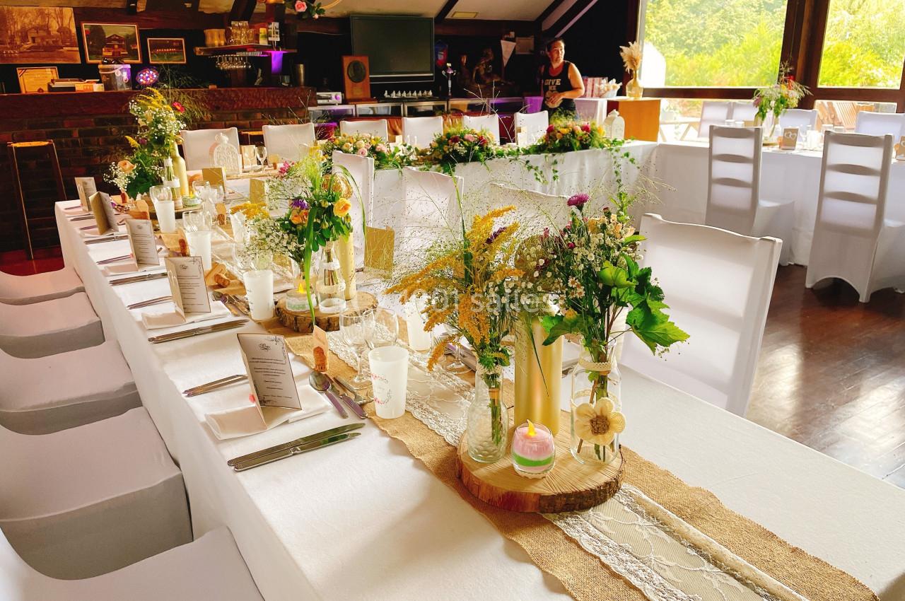 Table décorée pour un événement avec nappes blanches, fleurs, bougies et menus, dans une salle lumineuse.