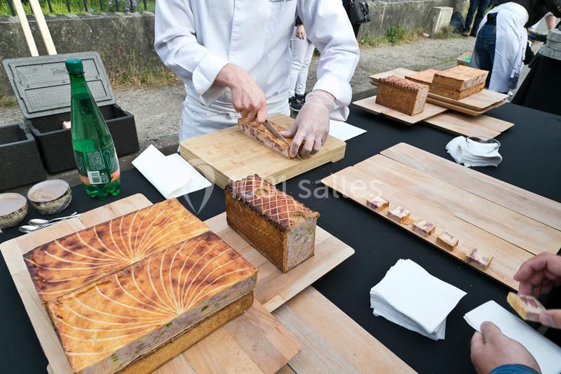 Un chef découpe un pâté en croûte sur une table en bois, entouré d'autres pâtés et d'ustensiles.