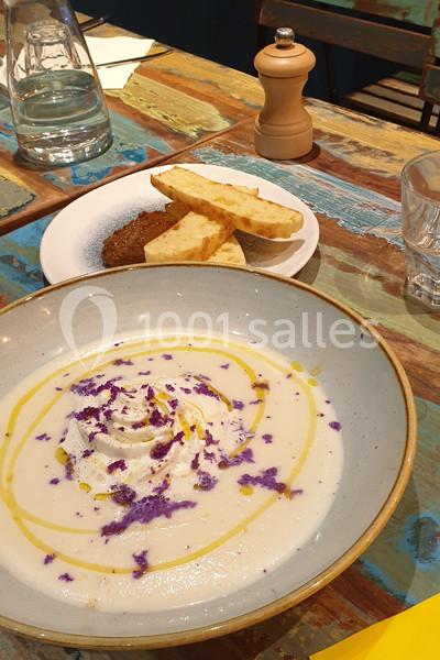 Bol de soupe crémeuse garnie de fleurs violettes, accompagné de pain grillé et d'une croquette sur une table colorée.