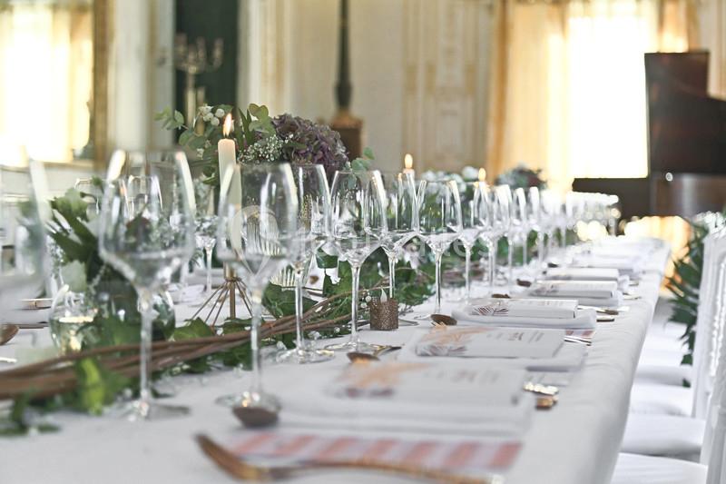 Table élégamment dressée avec nappes blanches, verres, couverts, bougies et décoration florale dans une salle lumineuse.