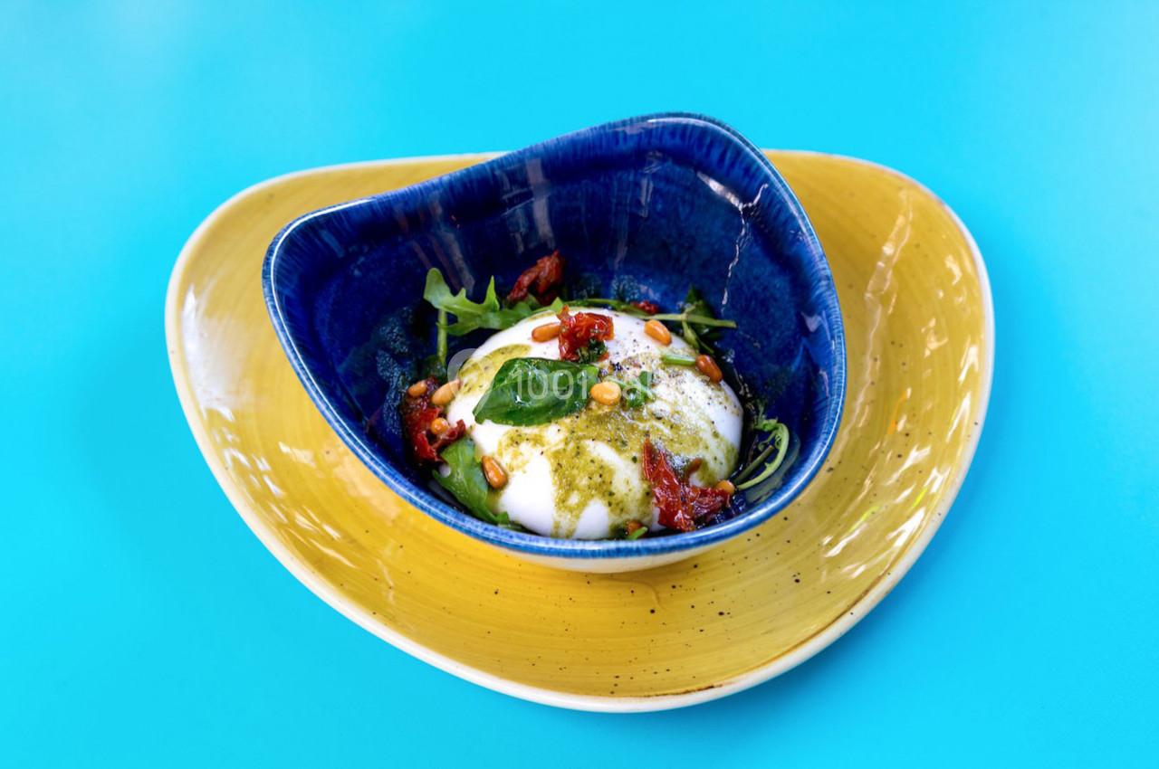 Assiette colorée contenant une burrata garnie de tomates séchées, pignons, roquette et pesto, sur fond bleu vif.