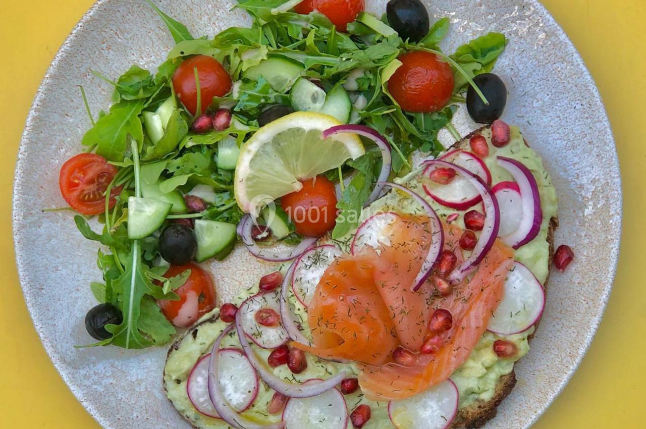 Assiette avec toast à l'avocat, saumon fumé, radis et grenade, accompagnée d'une salade verte, tomates et olives.