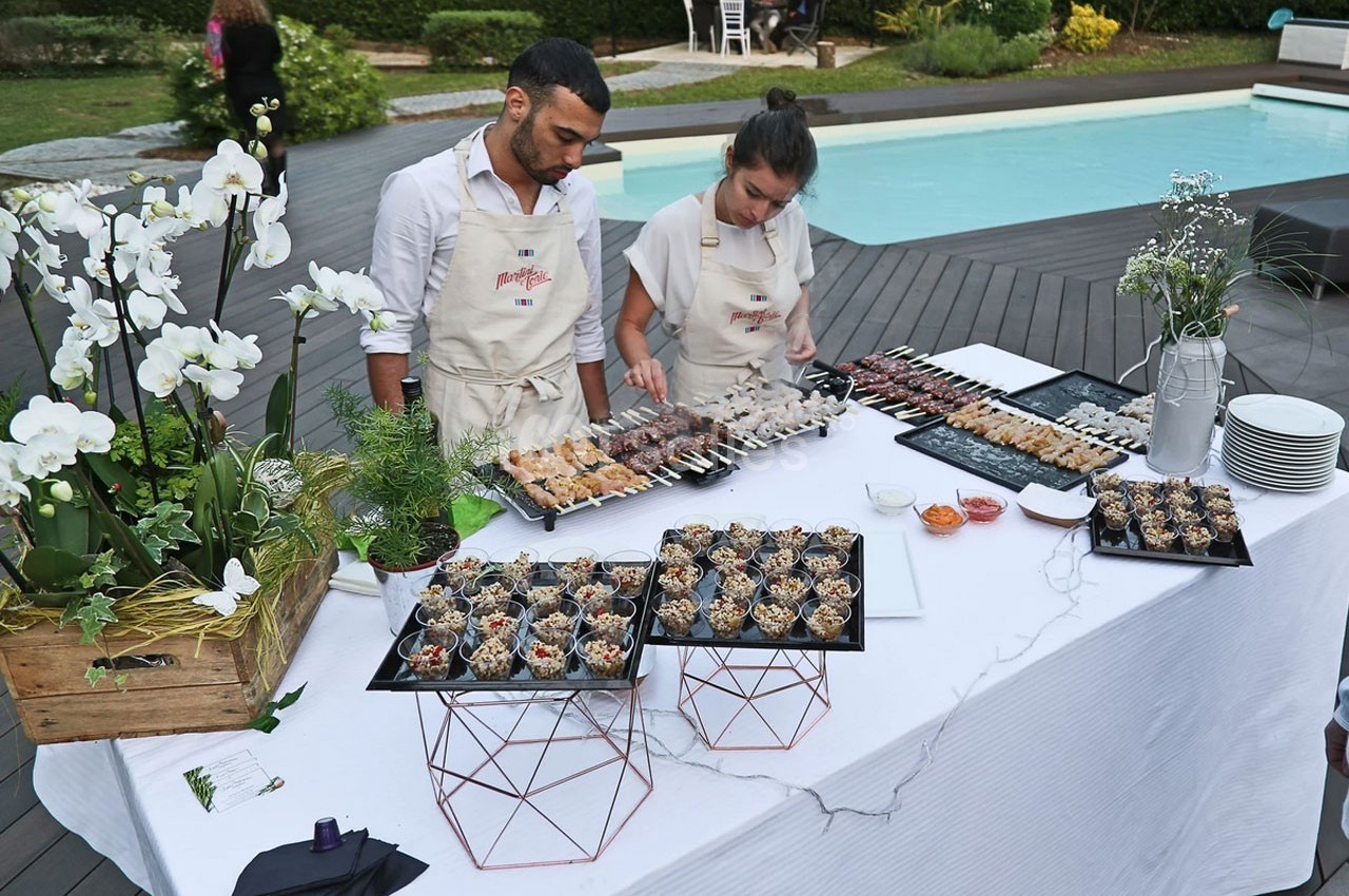 Deux personnes préparent un buffet de brochettes et verrines sur une table près d'une piscine dans un jardin.