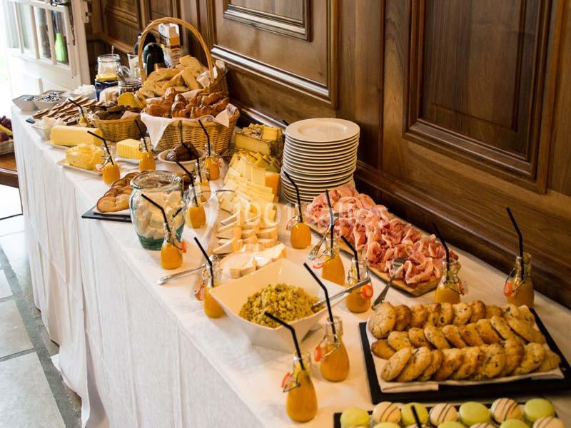 Buffet varié avec charcuterie, fromages, pains, viennoiseries, fruits et boissons disposés sur une table blanche.