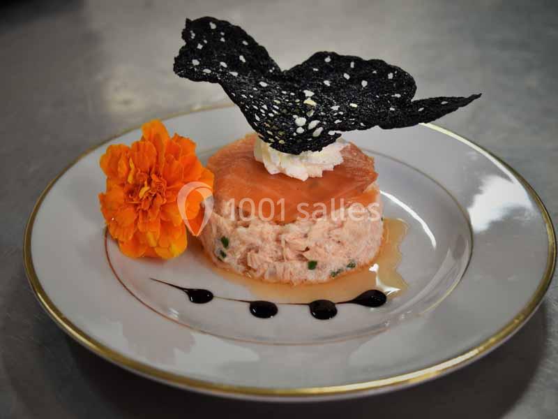 Tartare de saumon avec crème, tuile noire, fleur comestible et sauce décorative sur une assiette blanche.