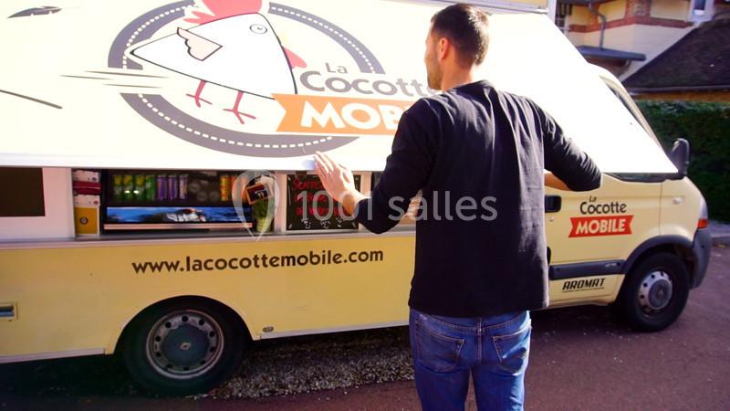 Un homme de dos interagit avec un food truck jaune stationné, affichant un logo de poule et des boissons visibles.