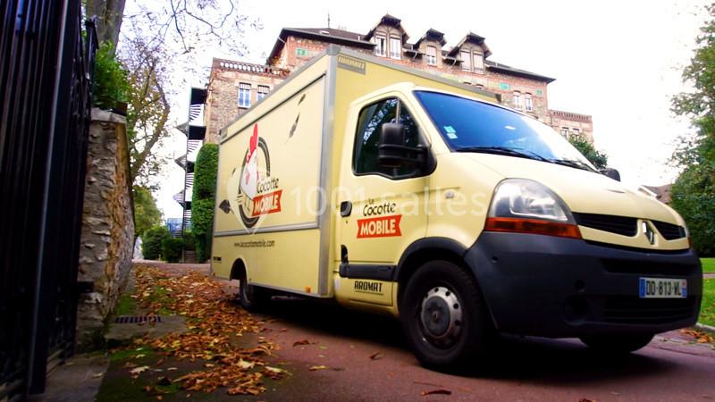 Camion utilitaire beige stationné sur une route bordée de feuilles mortes, devant un bâtiment en briques.