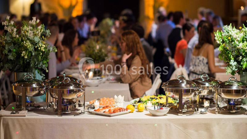 Buffet avec plats variés sur une table, entouré de convives assis à des tables décorées de fleurs.
