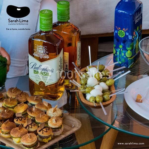 Plateau de mini-burgers, brochettes d'olives et mozzarella, avec bouteilles de boissons sur une table en verre.