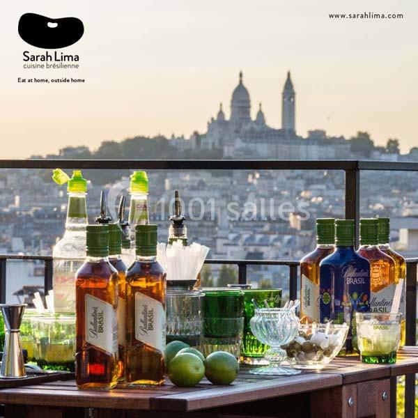 Table de bar en extérieur avec bouteilles, verres et citrons, vue sur Paris et la basilique du Sacré-Cœur en arrière-plan.