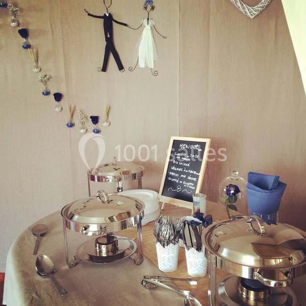 Buffet de mariage avec plats chauds sous cloches, couverts disposés et tableau de menu sur une table décorée.