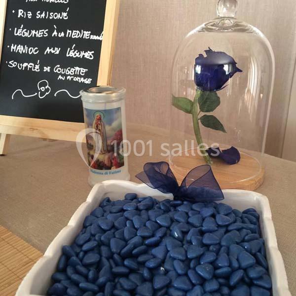 Une rose bleue sous cloche en verre, une bougie votive et des cailloux bleus dans un plat devant un tableau noir.