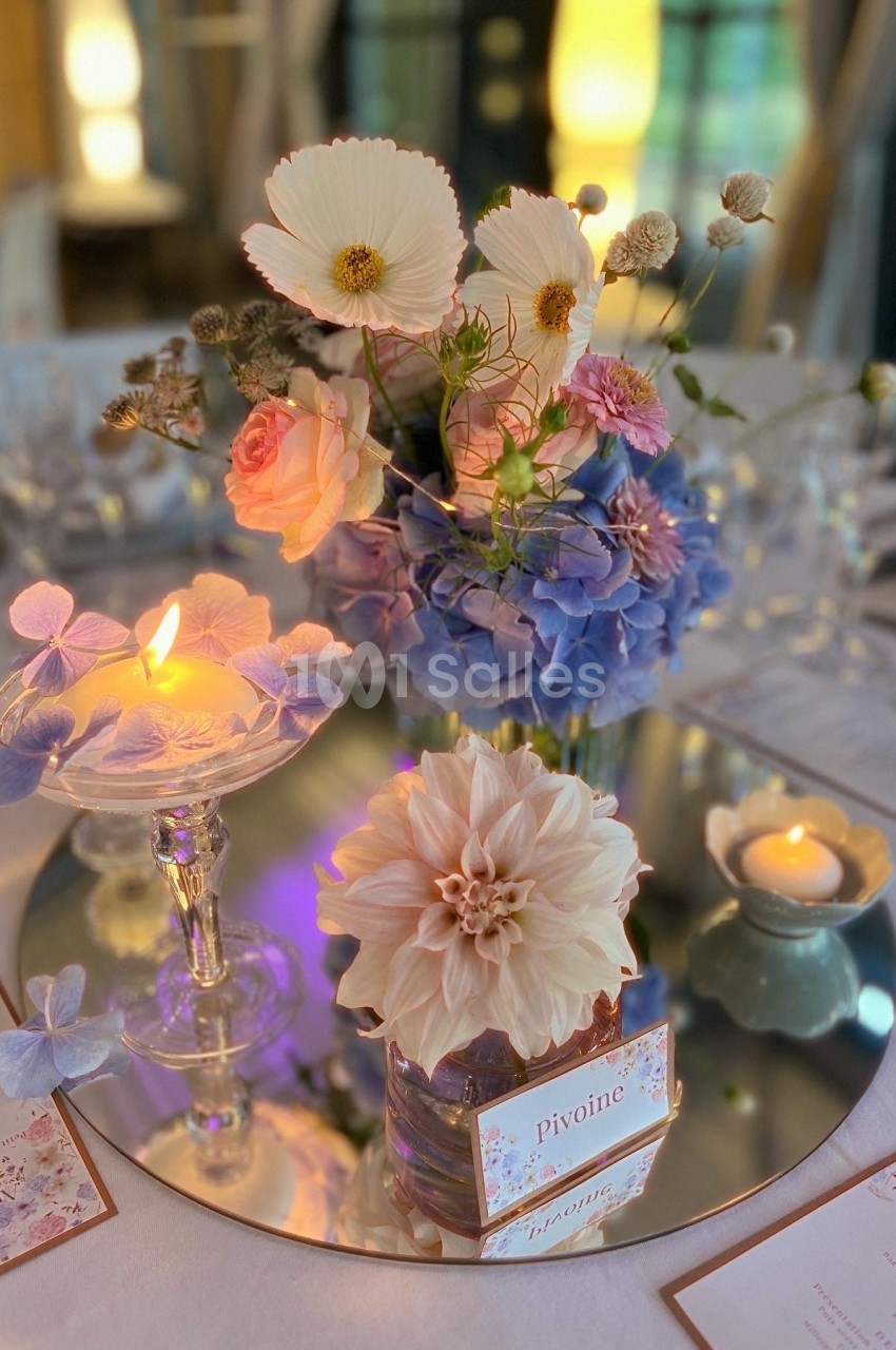 Centre de table floral avec dahlias, hortensias et cosmos, bougies allumées et menus disposés sur une table élégante.