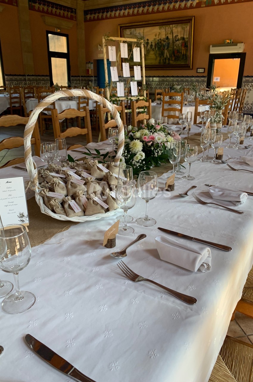 Table de réception décorée avec une nappe blanche, des fleurs, des couverts alignés et un panier de cadeaux invités.
