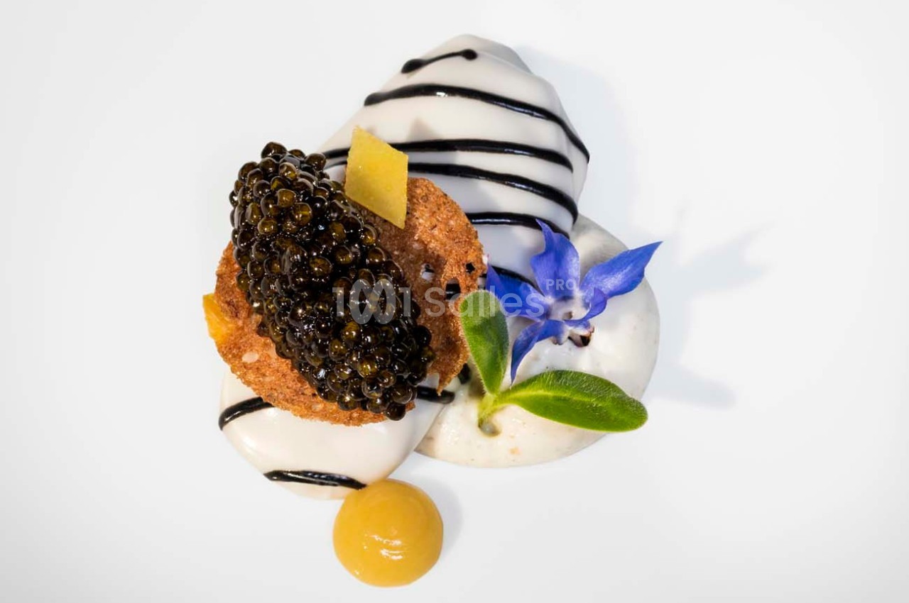 Dessert raffiné avec mousse blanche, caviar, biscuit, fleur comestible bleue et sauce jaune sur assiette blanche.