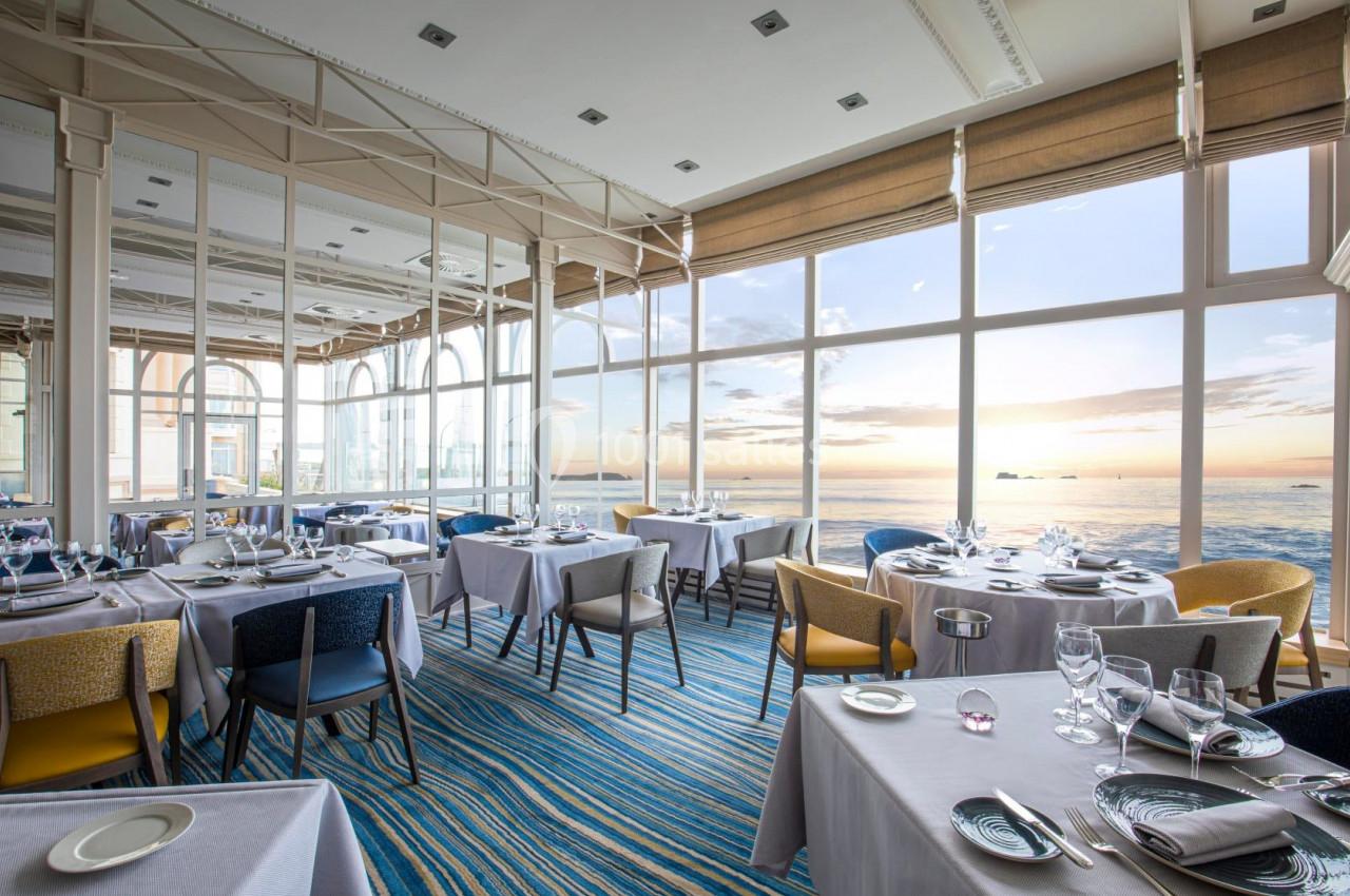 Salle de restaurant lumineuse avec grandes baies vitrées offrant une vue sur la mer au coucher du soleil.