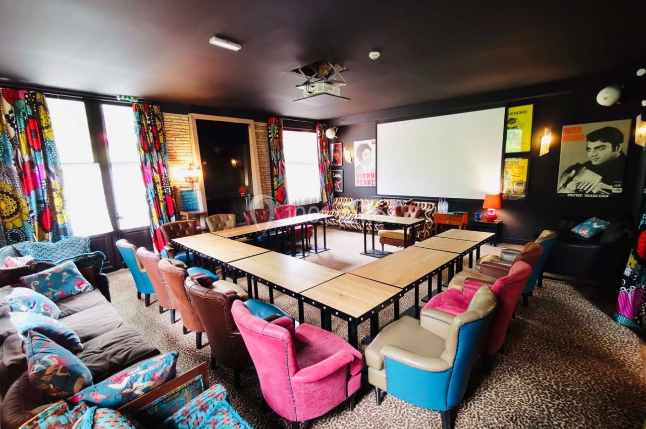 Salle de réunion colorée avec tables disposées en U, fauteuils variés, écran de projection et décorations murales.