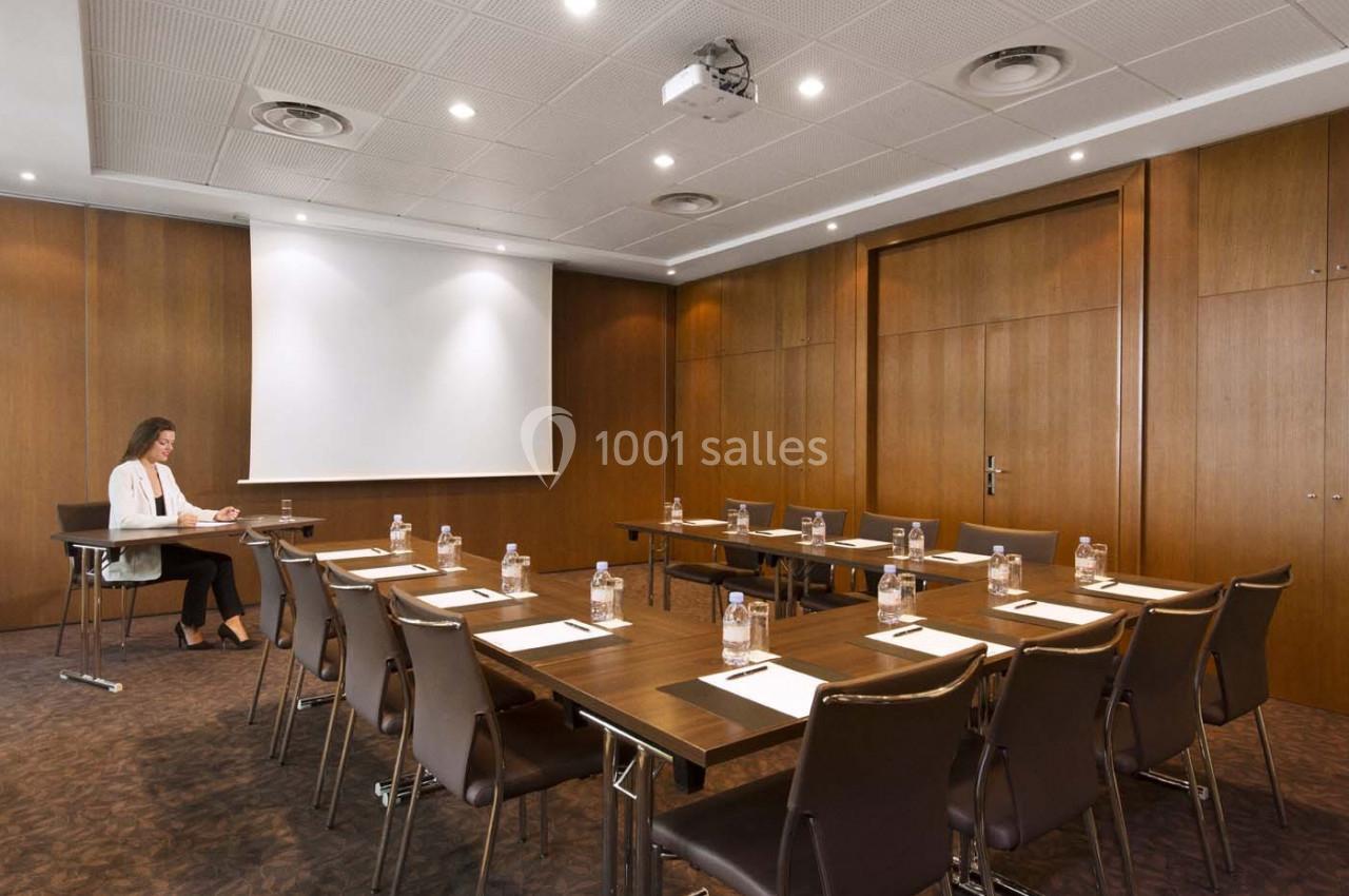Salle de réunion avec tables disposées en U, chaises, bouteilles d'eau, blocs-notes et une personne assise à gauche.