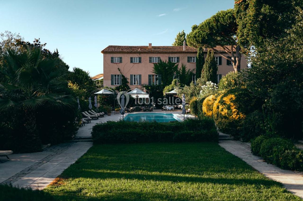 Grande maison en pierre entourée de jardins verdoyants avec une piscine centrale et des chaises longues.