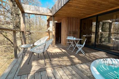 Terrasse en bois avec jacuzzi, deux fauteuils et table, offrant une vue sur un paysage verdoyant.
