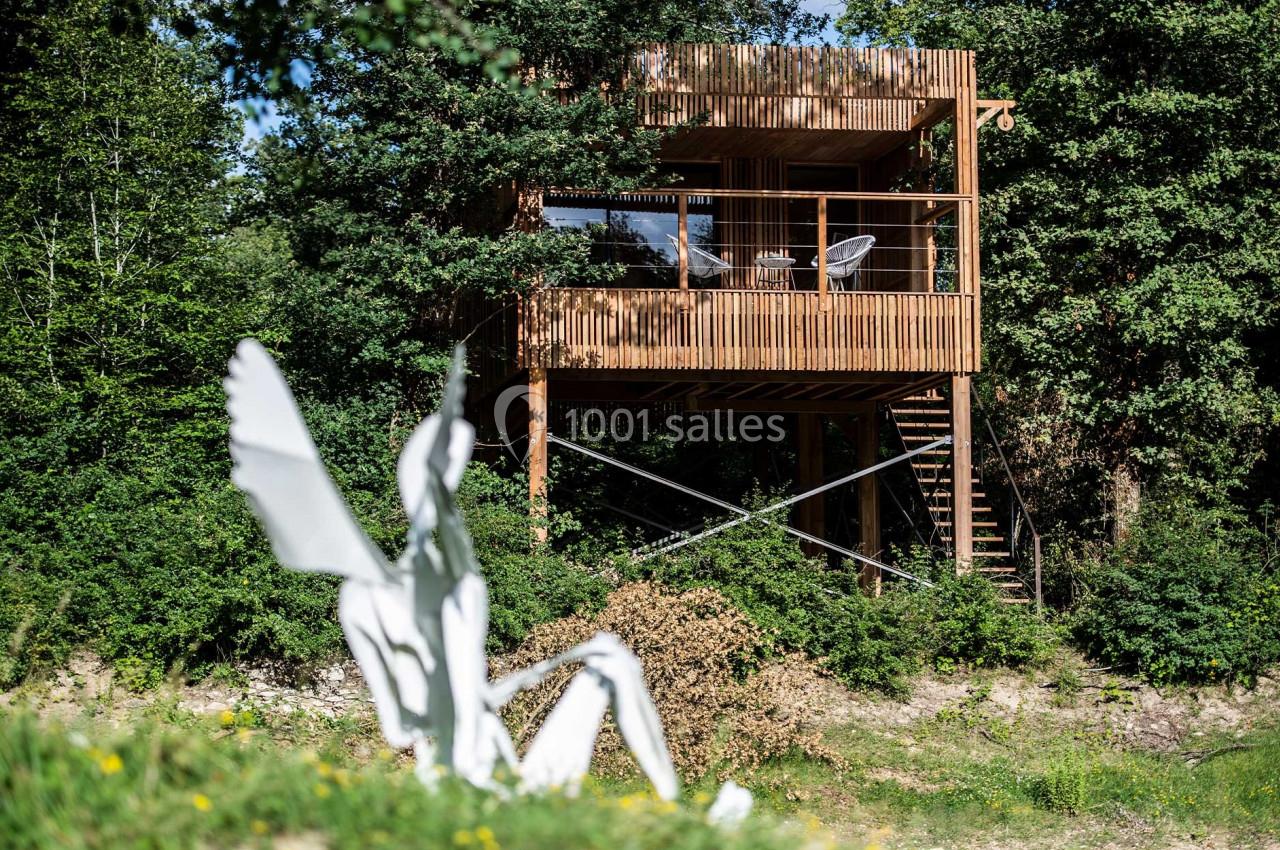 Cabane en bois sur pilotis entourée d'arbres, avec une sculpture blanche au premier plan dans un environnement naturel.