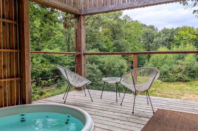 Terrasse en bois avec jacuzzi, deux fauteuils et table, offrant une vue sur un paysage verdoyant.