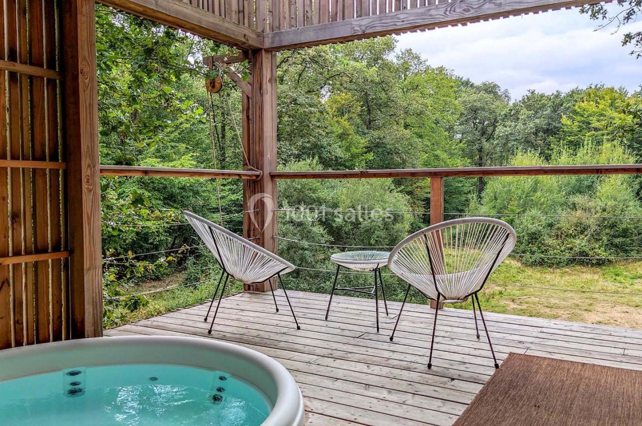 Terrasse en bois avec jacuzzi, deux fauteuils et table, offrant une vue sur un paysage verdoyant.