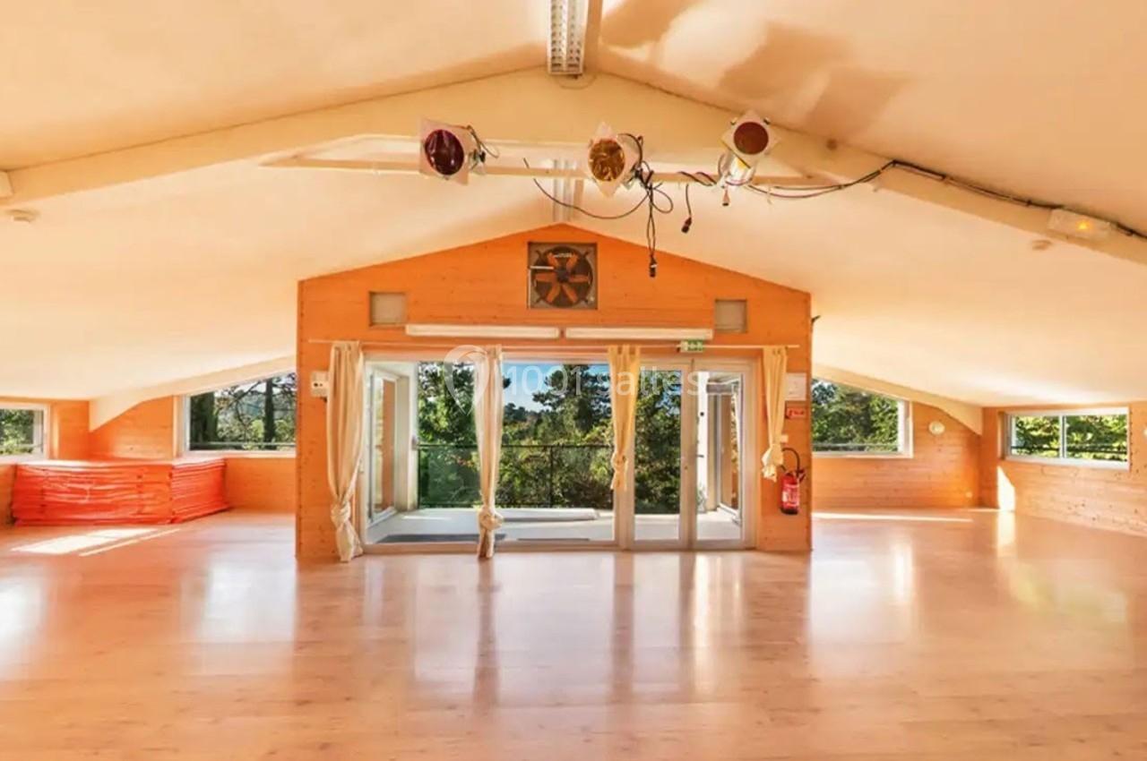 Salle lumineuse avec parquet en bois, grandes baies vitrées donnant sur un paysage arboré, et matériel rangé sur le côté.