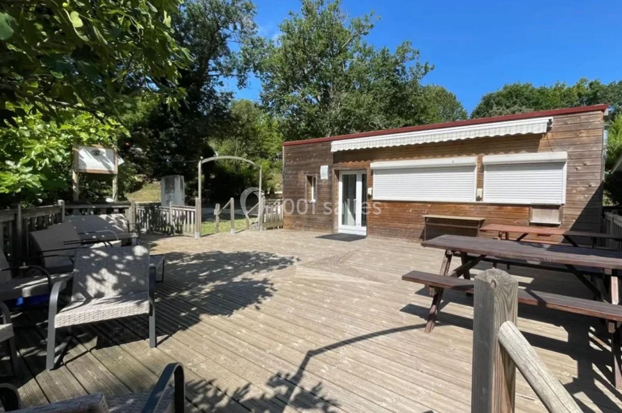 Terrasse en bois avec tables de pique-nique, chaises et bâtiment en bois sous un ciel bleu, entourée de végétation.