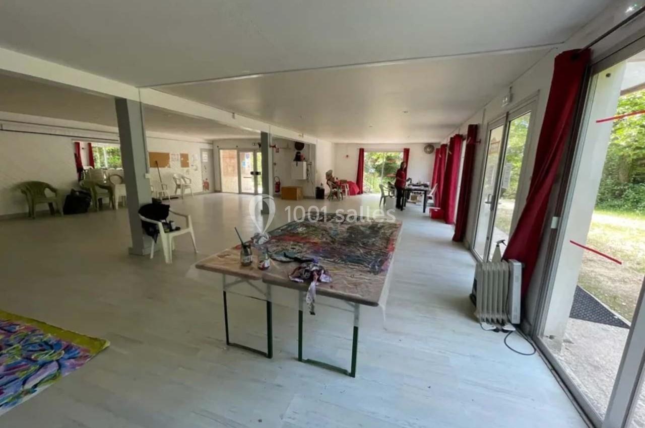 Salle lumineuse avec grandes baies vitrées, tables de travail, chaises empilées et matériel artistique dispersé.