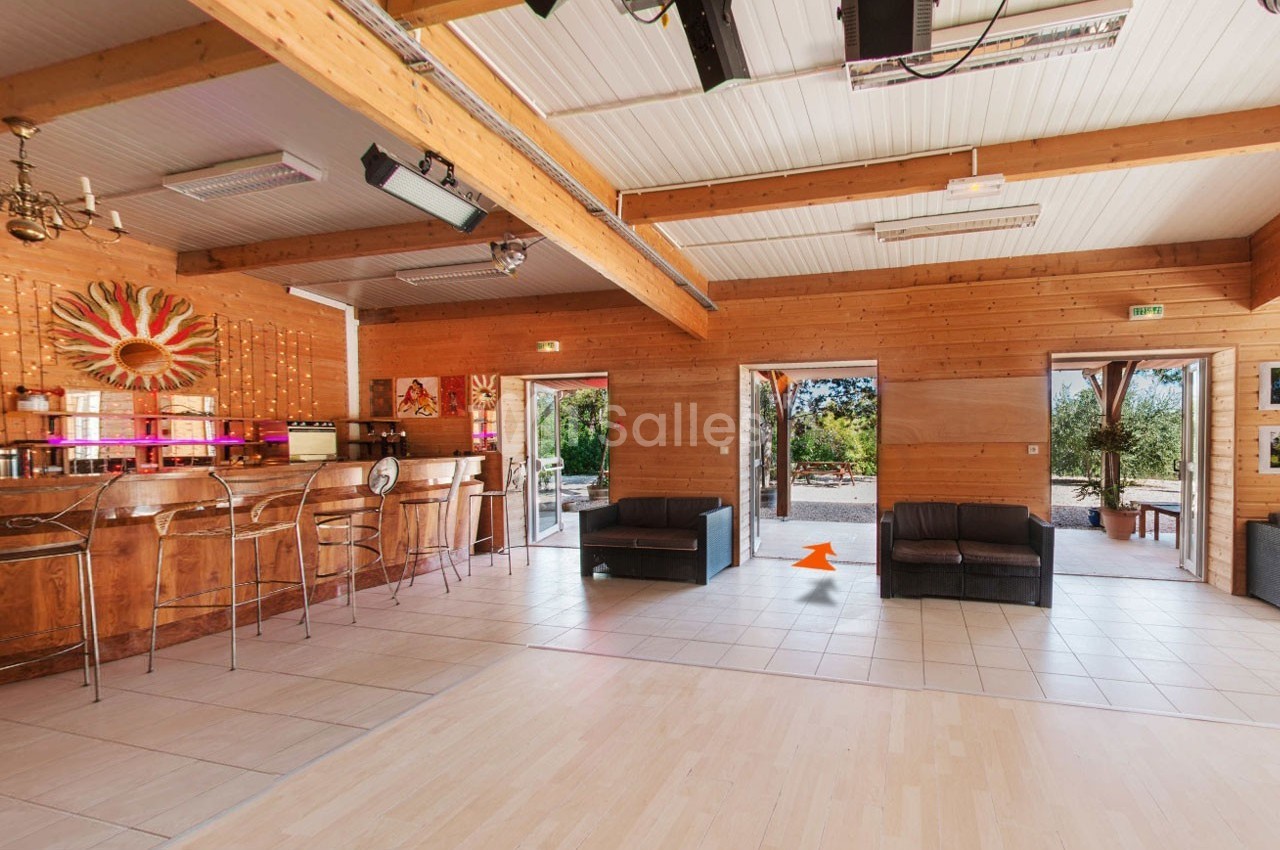Salle lumineuse avec murs en bois, bar équipé, canapés noirs et grandes ouvertures donnant sur un espace extérieur.