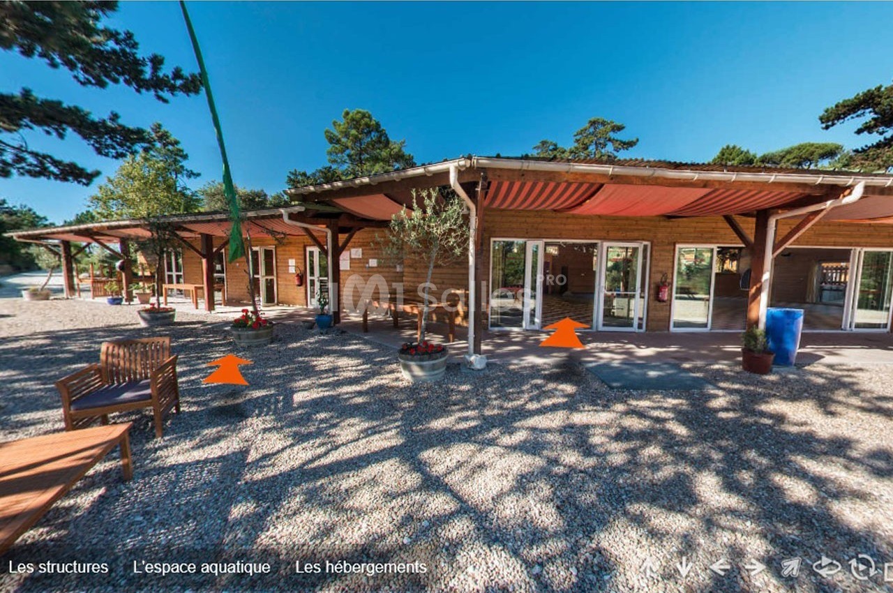 Entrée d'un bâtiment en bois avec terrasse ombragée, entourée de gravier et de végétation sous un ciel dégagé.