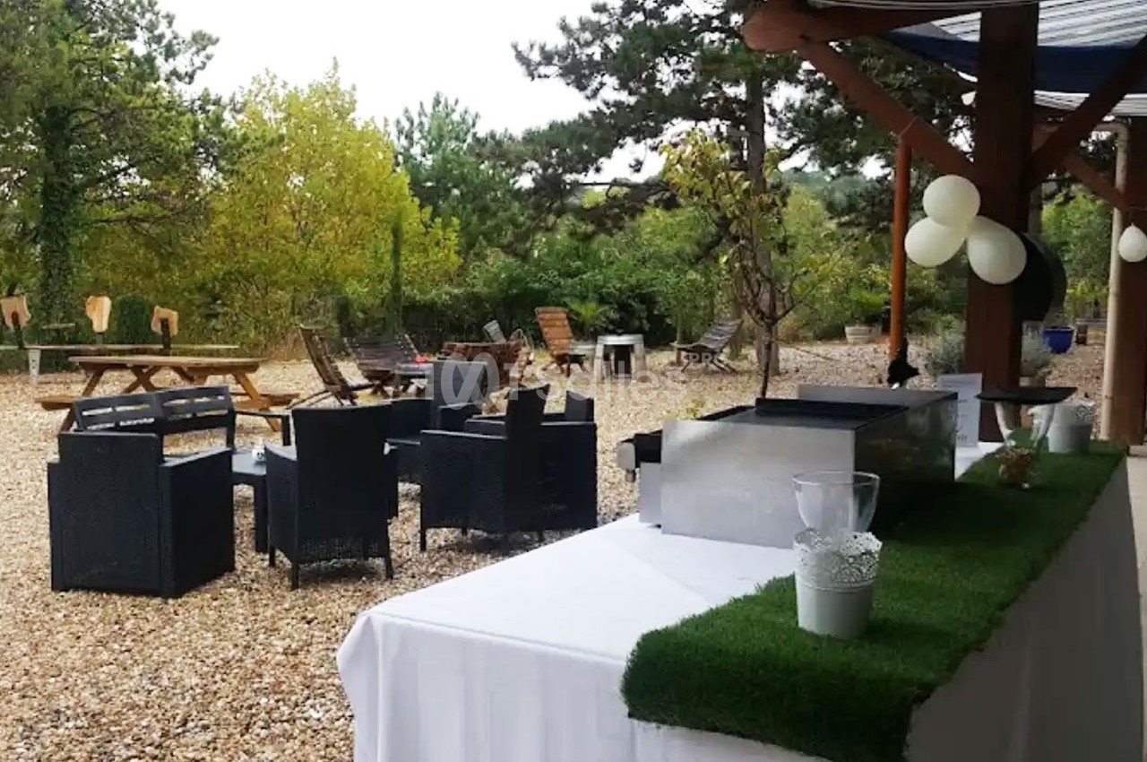 Espace extérieur avec mobilier de jardin, table de buffet et décorations, entouré de végétation et de graviers.
