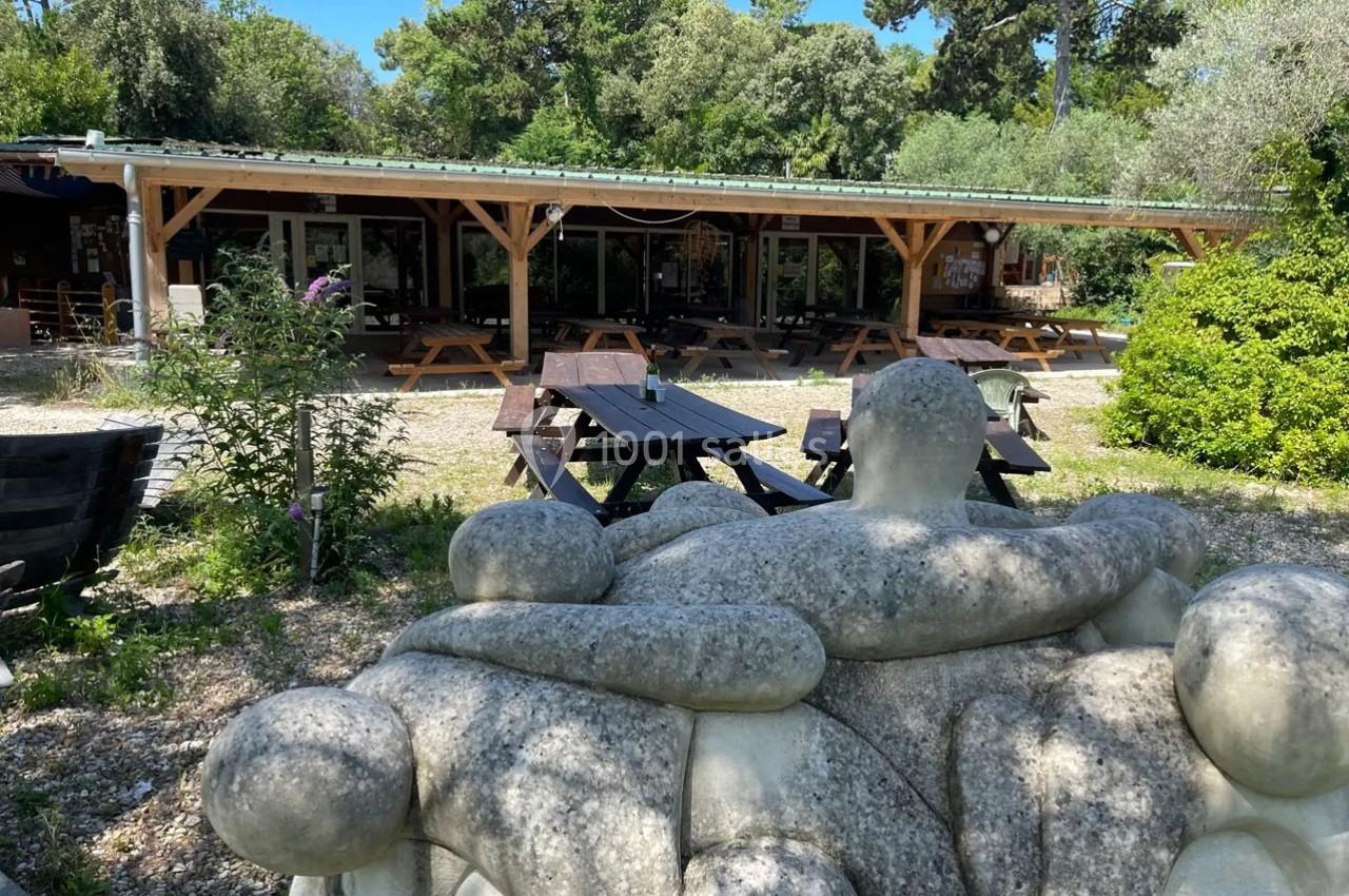 Statue en pierre représentant des figures humaines enlacées devant une terrasse en bois entourée de végétation.