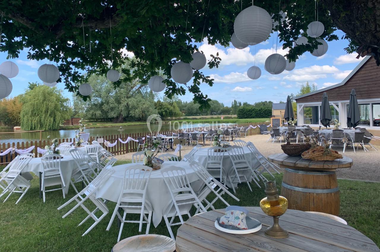 Tables dressées sous un arbre avec des lanternes suspendues, près d'un étang et d'une terrasse ensoleillée.