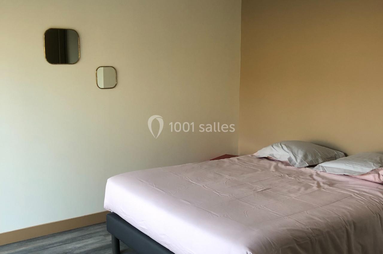 Chambre avec un lit double aux draps roses, sol en parquet gris, murs beige clair et suspension en métal décorative.
