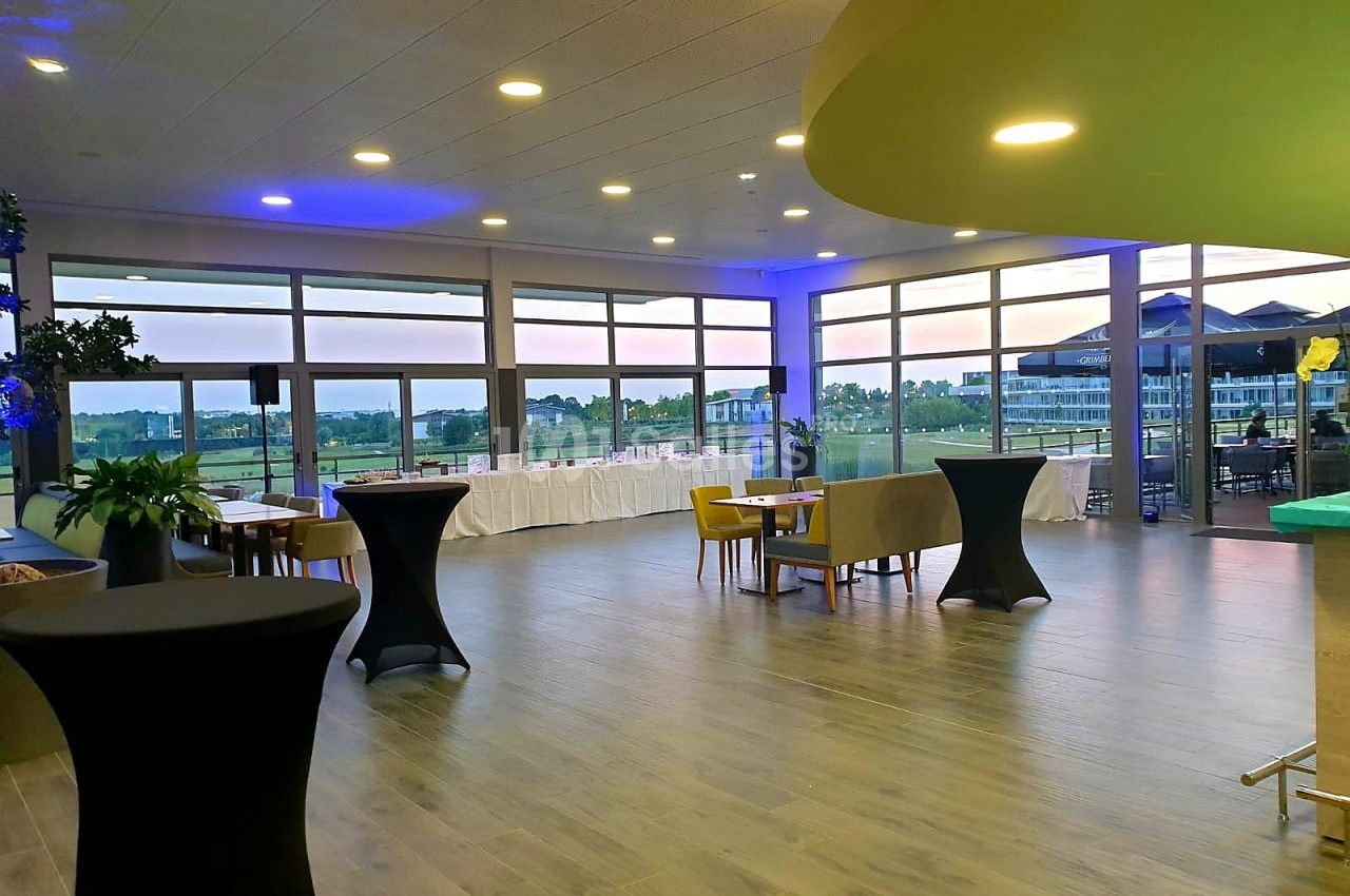 Salle lumineuse avec grandes baies vitrées, tables espacées et vue sur un paysage extérieur au crépuscule.