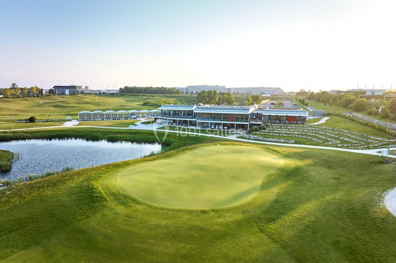 Vue d'un terrain de golf avec un green au premier plan, un étang et un bâtiment moderne en arrière-plan.