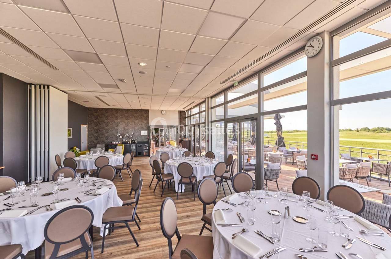 Salle lumineuse avec tables rondes dressées, grandes baies vitrées donnant sur une terrasse et un paysage verdoyant.